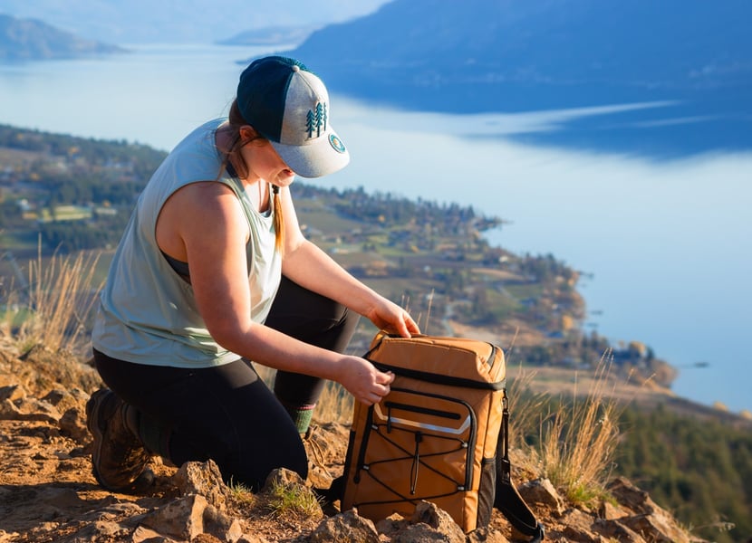 Woman kneeling in front of a cooler backpack on top of Spion Kop in Lake Country.