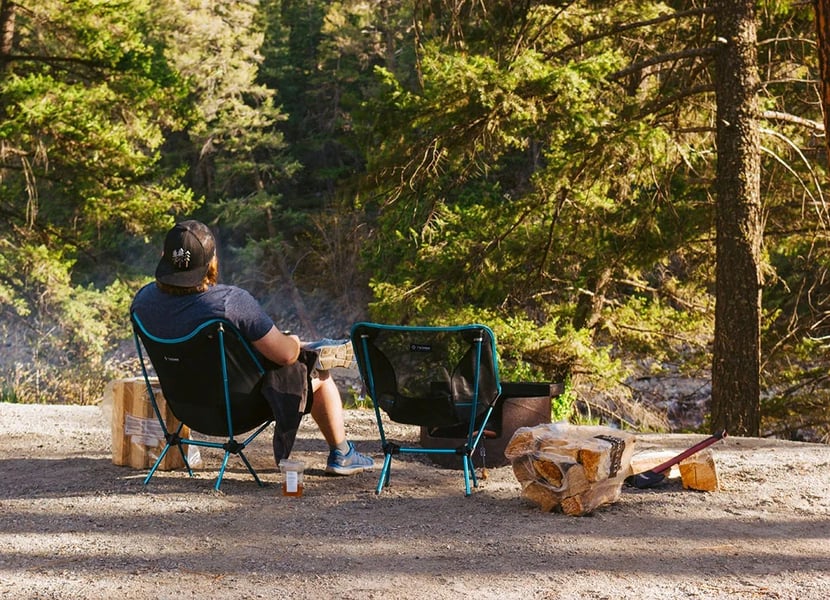 Man sits in a camping chair next to a campfire on a sunny day