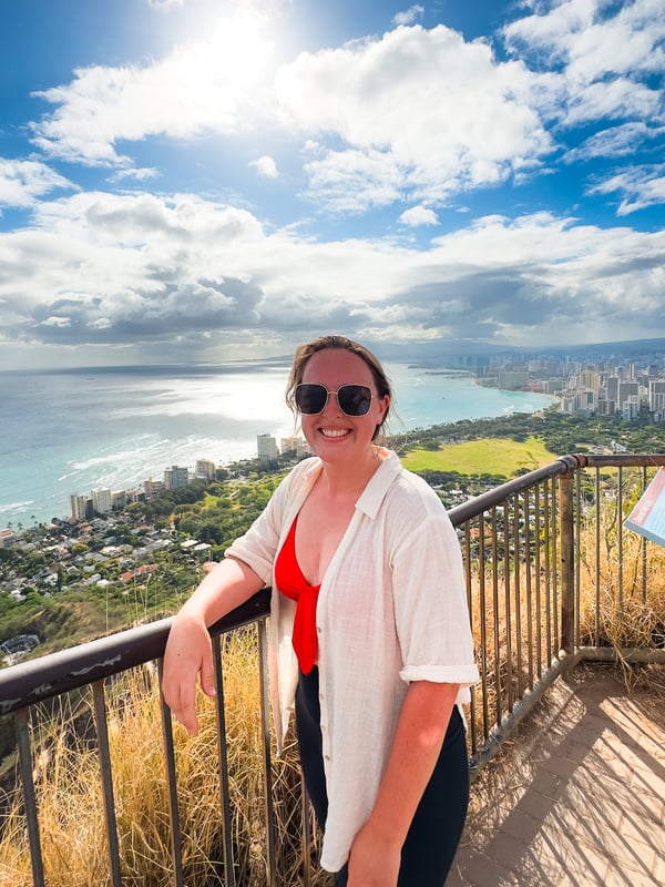 Woman leads against a guardrail at the summit of Diamond Head. Waikiki and the Pacific Ocean are in the background.
