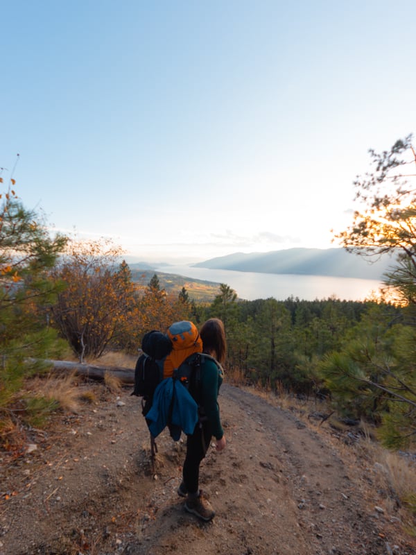 A woman with a heavy backpack looks over Okanagan lake at sunset.