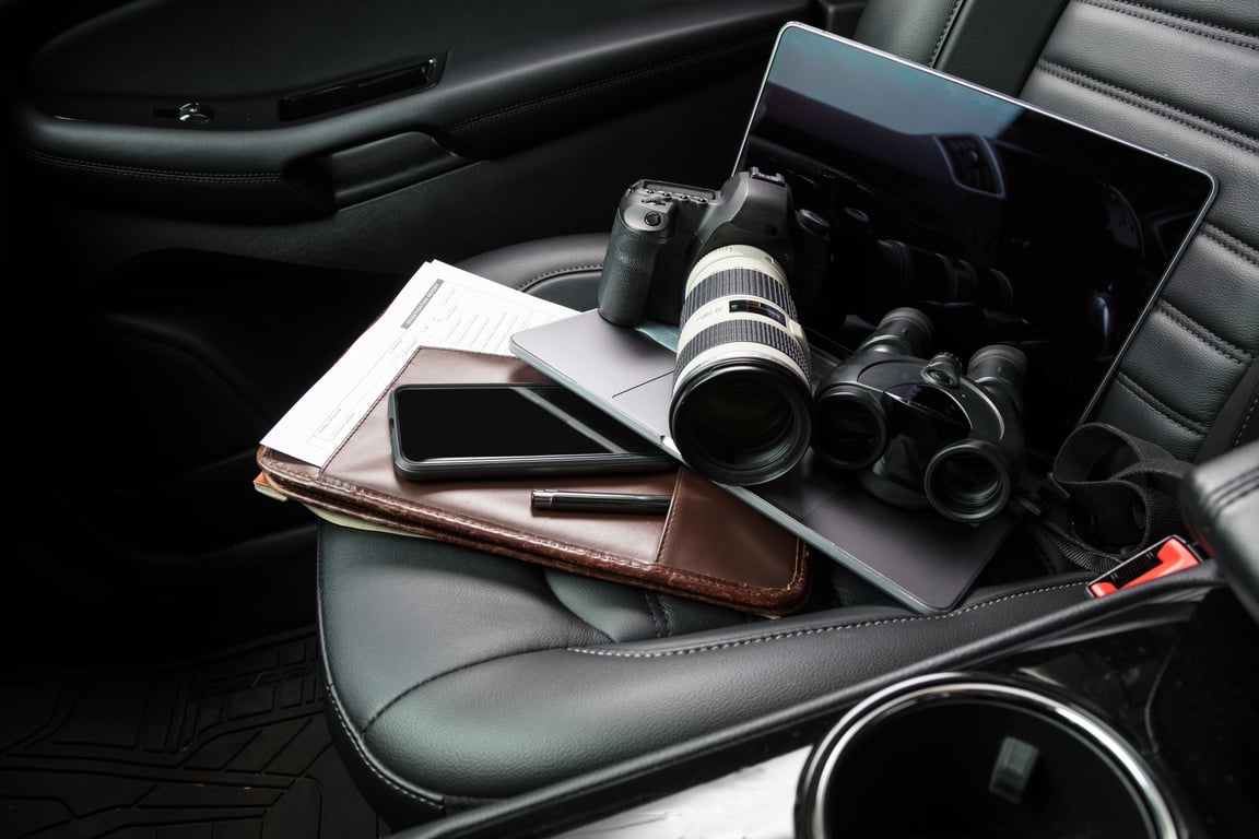 A collection of investigations equipment, including a camera with a telephoto lens, a tablet, and a notebook placed on a car seat.