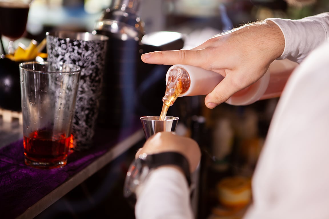 A close-up of a bartender's arms pouring a shot of brown alcohol