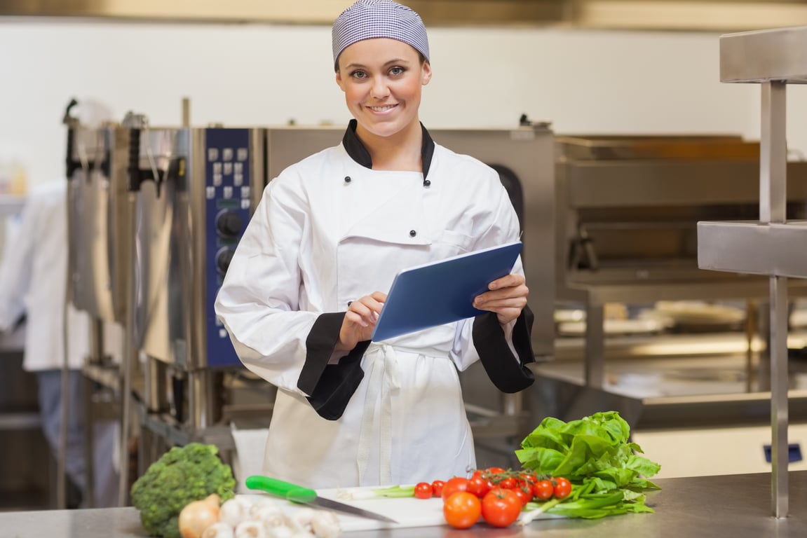 A smiling restaurant worker holds a clipboard while looking over fresh produce