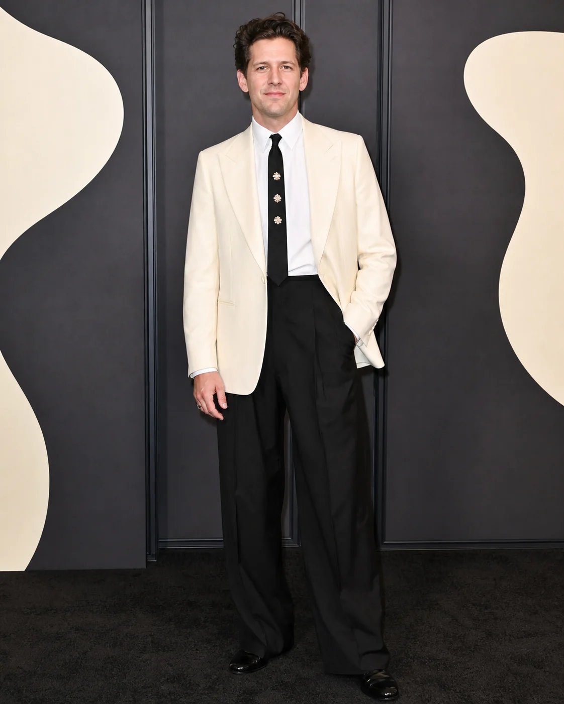 A man in a cream dinner jacket and brooch tie posing at a dark abstract press wall
