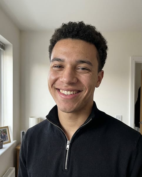 {
  "model": "NanoBanana",
  "task": "ultra realistic casual phone snapshot",
  "goal": "generate a highly natural, believable everyday portrait with zero studio feeling",
  "subject": {
    "person": "young man",
    "expression": "slightly asymmetric wide smile showing teeth, one lip corner raised a little higher, cheeks lifted with uneven natural tension, subtle eye squint, genuine candid joyful expression",
    "pose": "standing casually facing the camera, head tilted slightly a few degrees, shoulders relaxed, natural unstaged body language"
  },
  "appearance": {
    "hair": "short tight curly dark hair, natural shape, slightly imperfect everyday grooming",
    "skin": "visible pores, natural skin micro texture, subtle skin shine, slight tone variation, realistic facial imperfections, no retouching"
  },
  "clothing": {
    "top": "black quarter-zip sweater with silver zipper, simple casual everyday wear"
  },
  "environment": {
    "location": "ordinary indoor home environment",
    "background": "light indoor wall with subtle texture, faint shadow gradient, slight natural room depth, simple real-life setting",
    "context": "quick casual phone photo taken by a friend during a normal conversation"
  },
  "lighting": {
    "type": "natural mixed indoor light",
    "primary_source": "daylight coming from a nearby window on one side",
    "secondary_source": "soft ambient room light filling the opposite side",
    "effect": "slightly uneven illumination, soft natural shadow transitions, realistic exposure variation typical of phone photography"
  },
  "camera": {
    "device": "smartphone camera",
    "capture_style": "casual amateur phone snapshot",
    "lens_equivalent": "26mm smartphone lens",
    "framing": "head and shoulders portrait",
    "angle": "eye level",
    "focus": "natural phone autofocus on face",
    "depth_of_field": "natural smartphone depth",
    "artifacts": {
      "slight_noise": true,
      "subtle_compression": true,
      "minimal_lens_distortion": true,
      "slightly_compressed_dynamic_range": true,
      "minor_imperfection_in_sharpness": true
    }
  },
  "rendering": {
    "photorealism": "maximum",
    "style": "real everyday phone photography",
    "image_quality": "natural, candid, unpolished, believable",
    "detail": "high facial realism without looking processed or commercial"
  },
  "negative": [
    "studio portrait",
    "professional photoshoot",
    "beauty lighting",
    "perfect skin retouch",
    "plastic skin",
    "ai smile",
    "perfect symmetry",
    "cgi look",
    "cartoon style",
    "over smoothing",
    "commercial headshot",
    "over sharpened face",
    "harsh key light",
    "clean seamless backdrop",
    "fashion editorial look",
    "glamour portrait"
  ]
}