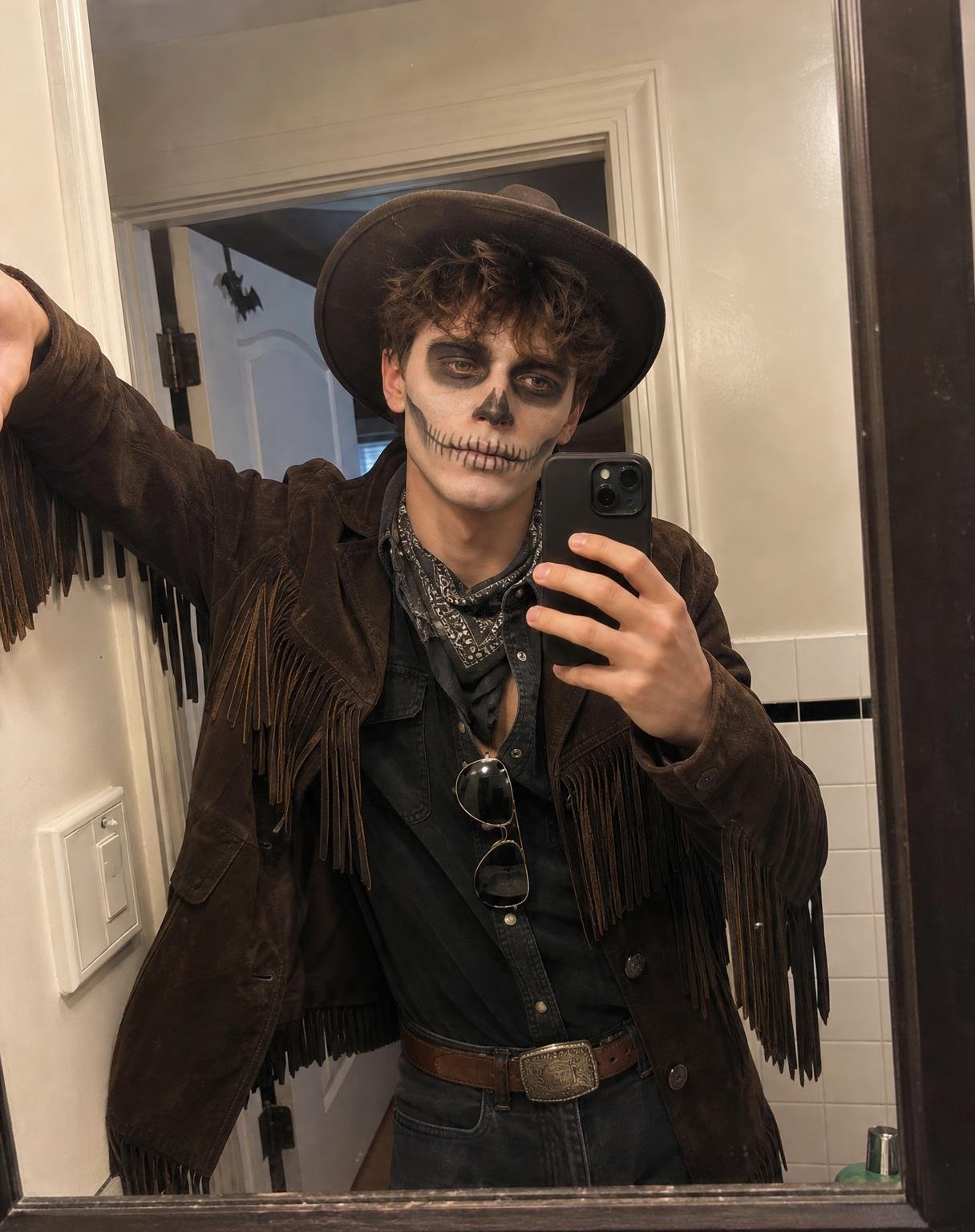 SKELETON COWBOY HALLOWEEN MIRROR SELFIE — ULTRA-REALISTIC 4:5
Create an extremely realistic, casual smartphone mirror selfie of a young man dressed as a skeleton cowboy for a Halloween party.
ABSOLUTE PRIORITY — FACIAL IDENTITY
Use the uploaded photograph of the young man strictly as his facial identity reference.
Preserve his recognizable facial structure, natural proportions, curly brown hair, forehead, eyes, nose, lips, jawline, skin tone, and overall appearance.
He must remain the same recognizable person underneath the Halloween makeup.
The reference photograph determines ONLY his identity. Completely replace the original outfit, outdoor café background, pose, lighting, and camera composition.
HALLOWEEN COSTUME — SKELETON COWBOY
Dress him in an authentic, slightly worn dark brown suede cowboy jacket featuring long, naturally hanging fringe across the chest, shoulders, and sleeves.
Underneath, he wears a dark charcoal button-down shirt with the top two buttons undone, faded black jeans, a worn brown leather belt with a silver western buckle, and a black-and-grey patterned bandana loosely tied around his neck.
Add a dark brown cowboy hat with a naturally curved brim, sitting comfortably over his messy curly hair.
The costume must look like real clothing someone assembled for a Halloween party, not a professionally styled fashion editorial.
REALISTIC SKELETON HALLOWEEN MAKEUP
Apply convincing, hand-painted skeleton makeup directly onto his face.
* Pale greyish-white face paint with slight unevenness and natural skin texture showing through.
* Deep black makeup around both eyes, following the natural contours of his eye sockets.
* A small black skeletal nose shape.
* Thin black skeletal teeth painted across his natural lips, extending slightly onto his cheeks.
* Subtle grey shading around the cheekbones and jaw.
* Slightly imperfect brushwork, uneven paint coverage, and authentic makeup texture.
The makeup should resemble a real Halloween costume created with ordinary face paint, not a digitally generated skull or a 3D skeleton mask.
Keep his natural facial anatomy visible beneath the makeup.
Do not add sunglasses over his eyes. A pair of aviator sunglasses can hang casually from his shirt collar.
MIRROR SELFIE — COMPOSITION
Photograph him taking a casual mirror selfie inside an ordinary residential bathroom.
Use a large, nearly square rectangular wall-mounted mirror, similar to the supplied Halloween cowboy inspiration image.
The mirror has a simple dark frame with realistic thickness and slightly imperfect edges.
Do not use a tall, narrow, full-length bedroom mirror.
The man stands slightly off-centre, leaning casually against the bathroom doorway.
One arm is raised and rests loosely against the doorframe, with his elbow bent. His other hand holds a dark smartphone in front of his upper chest.
The smartphone must have a realistic size, correct camera placement, and anatomically accurate fingers.
His face and skull makeup remain clearly visible.
Frame the reflection from the top of his cowboy hat to approximately his upper thighs.
The composition should feel slightly imperfect and naturally cropped, like a real person quickly taking a Halloween costume selfie before leaving for a party.
BATHROOM ENVIRONMENT
Create a believable, modest residential bathroom with off-white painted walls, an ordinary doorframe, a small section of tiled wall, and subtle everyday details.
The bathroom should look genuinely lived-in rather than professionally decorated.
A slightly open door reveals a small portion of an adjoining room.
Include only minimal, believable Halloween decoration, such as a small paper bat decoration near the doorway.
The costume and skull makeup should immediately communicate Halloween without excessive decorations.
Maintain physically accurate mirror reflections and realistic room geometry.
LIGHTING — MOST IMPORTANT
Use ordinary indoor residential lighting, similar to a casual smartphone photograph taken in the evening.
The light comes from a normal ceiling fixture, producing slightly uneven illumination across his face and clothing.
Keep natural shadows around his eyes, underneath the cowboy hat, and along the bathroom doorway.
Avoid bright orange string lights, artificial golden lighting, cinematic illumination, glowing skin, studio lighting, or dramatic rim lights.
Do not artificially brighten the entire room.
Allow some areas of the background to appear naturally darker.
The photograph should have slightly imperfect exposure, realistic shadow noise, subtle smartphone grain, and ordinary indoor white balance.
PHOTOGRAPHIC REALISM
The final image must look like a genuine, unedited Halloween mirror selfie taken with an iPhone.
Preserve natural skin texture beneath the face paint, realistic hair strands, authentic suede fabric texture, natural jacket fringe movement, believable reflections, and correct human anatomy.
Avoid perfectly symmetrical posing, artificial facial smoothness, exaggerated sharpness, glossy surfaces, synthetic textures, and overly clean environments.
No beauty filters, HDR processing, cinematic colour grading, excessive background blur, or professional fashion photography aesthetics.
The photograph should feel ordinary, spontaneous, and human.
OUTPUT REQUIREMENTS
* Aspect ratio: 4:5 vertical.
* Natural upper-thigh framing.
* Nearly square mirror with realistic proportions.
* Clearly visible skeleton face makeup.
* Dark brown fringe cowboy jacket.
* Authentic cowboy hat and Halloween styling.
* Realistic bathroom mirror reflection.
* Natural, imperfect indoor smartphone lighting.
* No text, logos, watermarks, borders, or collage.
FINAL PRIORITY: The result must resemble an authentic Halloween costume selfie someone casually posted on Pinterest or Instagram, not a polished AI-generated portrait. Prioritize photographic believability over beauty, perfection, or dramatic styling.