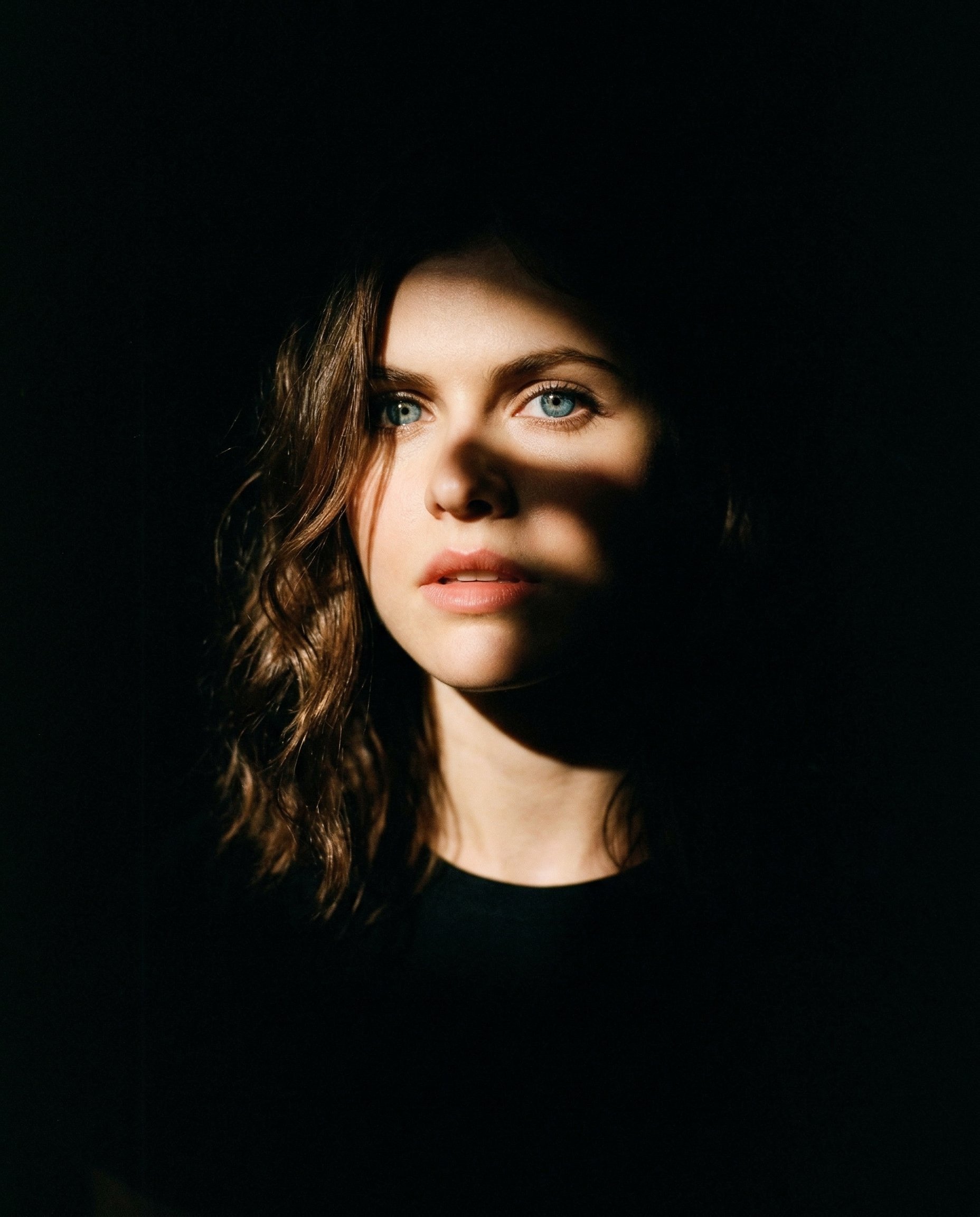 INPUT:
- Reference image of a person (must be used as identity base)

PROMPT:
Use the reference image as the exact identity base. Create a cinematic portrait with dramatic partial lighting, as if the subject is emerging from darkness with a strong, focused spotlight partially blocked by an object.

CORE LIGHTING (MOST IMPORTANT):
- Use a single hard spotlight coming from one side (slightly above eye level, angled)
- Simulate the light being **partially blocked by an object**, creating an irregular shadow cut across the face
- Only ONE side of the face should be illuminated (around one eye, cheek, and lips)
- The other side must fall into deep, pure black shadow
- The shadow edge must feel organic and imperfect (not a clean straight line)

LIGHT QUALITY:
- Strong, high contrast
- Bright highlight on skin where light hits
- Soft falloff into darkness
- No fill light — shadows must stay deep and rich

COMPOSITION:
- Slightly off-center framing (not perfectly symmetrical)
- Close portrait (head and shoulders)
- Natural angle, not stiff

FACE & EXPRESSION:
- Neutral or slightly intense expression
- Lips slightly parted (optional for cinematic feel)
- Eyes sharp and engaging

BACKGROUND:
- Completely dark or near-black
- No visible environment

STYLING:
- Hair softly lit on one side, fading into darkness on the other
- Optional subtle jewelry catching light (like earrings)
- Clothing dark, blending into shadows

COLOR GRADING:
- Warm cinematic tone on highlights (golden/orange tint)
- Deep crushed blacks
- Slight film-like contrast

TEXTURE:
- Add fine film grain
- Slight natural imperfection (not overly clean or digital)

EFFECTS:
- NO glow
- NO neon
- NO glitch
- NO overlays

DEPTH:
- Strong subject separation using light vs darkness only

MOOD:
- Cinematic, intimate, slightly mysterious
- Feels like a movie still or vintage film frame

IMPORTANT:
- Do NOT evenly light the face
- Do NOT center the light
- The asymmetry and shadow cut is the key visual hook