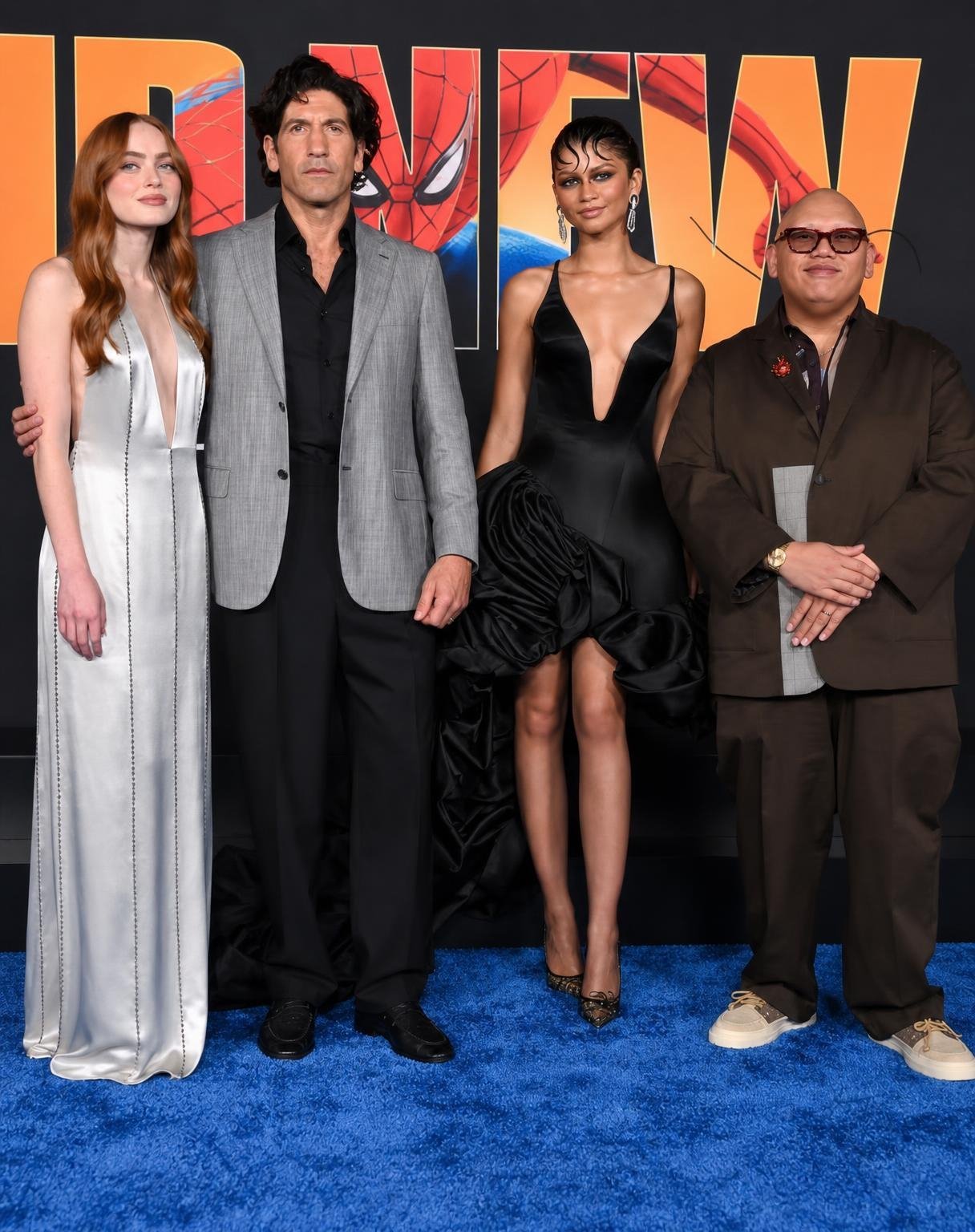 remove the man wearing the burgundy-red suit in the center of this photo completely. Bring the remaining four people closer together naturally to close the empty space. Reconstruct the background seamlessly. Preserve their original faces, outfits, expressions and poses. Make it look like the removed person was never in the photograph. No visible gaps, awkward spacing or editing artifacts.