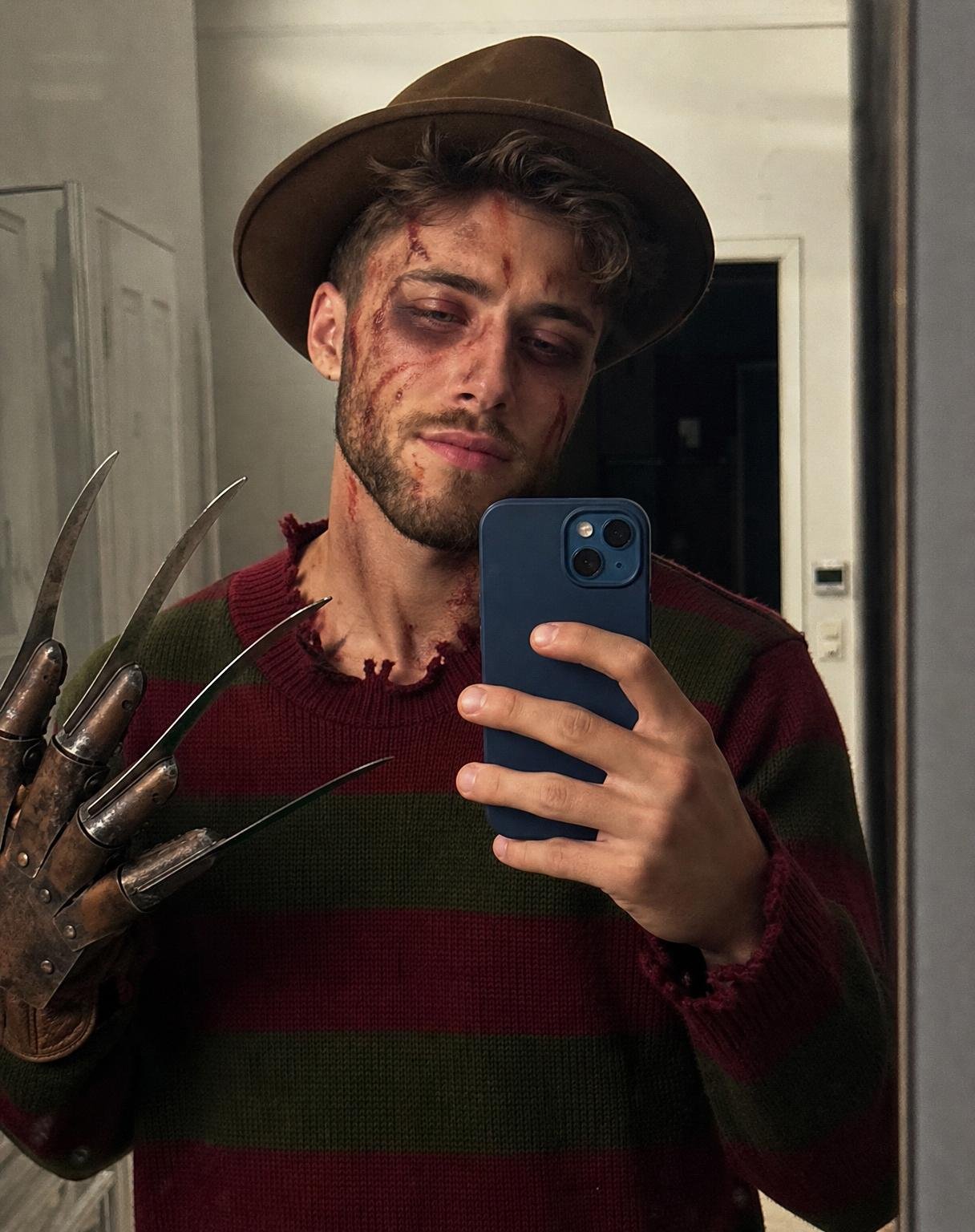 Generate a completely new, photorealistic iPhone mirror selfie of the person in the uploaded facial reference, dressed in a Freddy Krueger-inspired Halloween costume.
CRITICAL — NATURAL FACE, NOT FACE SWAP: Use the reference image only to understand the person’s identity. Generate a fresh, three-dimensional face naturally belonging to the photographed person. Do not copy, cut out, paste, overlay, or transplant the original face pixels. Preserve the person’s recognizable facial characteristics while naturally adapting their expression, head angle, perspective, and facial anatomy to the new selfie.
The face must be rendered as part of the original photograph, with naturally connected temples, ears, jawline, neck, and hairline. Match the lighting and focus across the entire head. Halloween makeup must follow the actual contours of the face, with realistic texture over the skin rather than appearing as a separate digital layer.
HALLOWEEN APPEARANCE: A tan vintage fedora, an oversized dark olive-green and burgundy striped knitted sweater, and dark pants. Add scary Freddy Krueger-inspired makeup with ashy pale skin, dark bruised eye sockets, irregular red scratch marks, realistic smudged bloody streaks, and subtle scar details. Keep it convincing as real Halloween makeup applied by hand.
POSE AND COMPOSITION: A casual, slightly tilted iPhone mirror selfie in a simple indoor hallway. Hold a blue dual-camera iPhone naturally in one hand, partially covering the lower face and chest. Raise the other hand beside the body, wearing a weathered brown leather horror glove with four long metallic blades. Frame from the hat to the upper thighs. Keep the same relaxed, casual mirror-selfie energy as a real personal photo.
LIGHTING: Ordinary overhead room lighting and natural phone-camera exposure. Realistic shadows beneath the hat brim, around the nose, under the chin, and along the neck. Natural reflections on the phone and glove. Consistent sharpness, grain, white balance, and color across the face, hair, clothing, and background.
REAL PHOTO QUALITY: Authentic iPhone mirror selfie, natural skin pores, slight asymmetry, imperfect hair strands, visible knit texture, subtle mirror dust, mild sensor grain, ordinary indoor colors, and unedited photographic realism. Avoid the glossy, overly smooth appearance of AI portraits.
DO NOT: Create a face-swap image, preserve the reference image’s original lighting, place a sharp face over a softer head, produce mismatched skin texture, blur the facial edges, create a floating face, beautify the identity, or make the result resemble a movie poster.
The final image should look as if the person genuinely dressed up for Halloween, stood in front of a mirror, and took one real photograph with their iPhone.