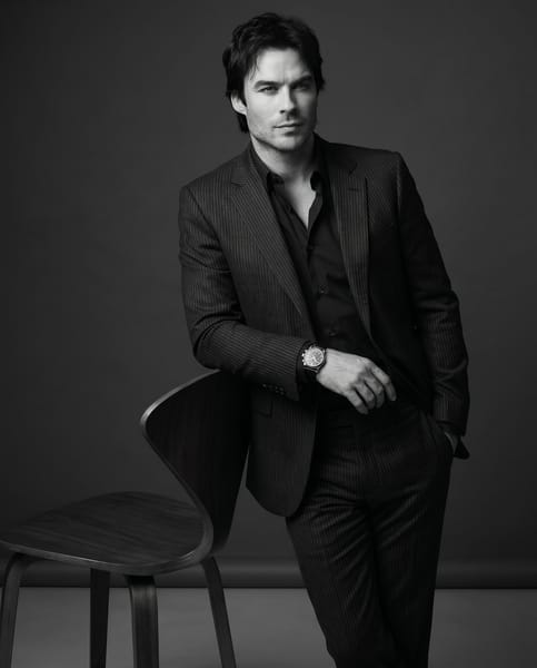 Create an ultra-photorealistic black-and-white fashion editorial portrait with a cinematic luxury studio aesthetic: elegant, minimal, refined, powerful, and magazine-quality.
Composition:
Medium-shot vertical 4:5 portrait.
The subject is slightly off-center, leaning confidently against a modern backward-facing wooden studio chair. The upper body, chair, both hands, wristwatch, and part of the lower body must be visible. Keep clean editorial framing, strong negative space, and no awkward cropping.

Pose:
The subject is not seated. They lean naturally against the chair in a relaxed but powerful posture. One arm rests over the top edge of the chair back, while the other hand stays naturally inside the trouser pocket. The wristwatch must be clearly visible. Shoulders relaxed, posture composed, body language strong, self-assured, and editorial.

Expression & Gaze:
Calm, confident, self-assured expression with closed lips or a subtle neutral look. The subject must not look sad, tired, weak, shy, dull, or emotionally heavy. Gaze slightly off-camera to the side, not directly into the lens.

Wardrobe:
Perfectly tailored slim-fit dark pinstripe suit with subtle vertical stripes, paired with a dark open-collar shirt. In black-and-white, the suit and shirt must have clear tonal separation: suit slightly lighter or more textured, shirt slightly deeper or smoother. The outfit should look sharp, structured, premium, fitted, with crisp tailoring lines and realistic fabric texture.

Hair & Styling:
Preserve the subject’s hairstyle and natural texture accurately. Styling should feel polished, elegant, and fashion-editorial. Keep makeup or grooming minimal and refined, enhancing the face without looking artificial.

Environment:
Minimal seamless dark grey gradient studio backdrop. No visible edges, corners, extra props, text, logos, or distractions except the wooden chair.

Lighting:
Moody directional studio lighting in pure monochrome. Use strong but controlled contrast with soft falloff to sculpt the face, cheekbones, jawline, hair, suit texture, shirt separation, wristwatch, hands, and chair details. Maintain cinematic depth, rich tonal range, and no harsh overexposure.

Quality & Style:
Ultra-realistic 8K monochrome fashion editorial photography. Crisp focus on the face, eyes, lips, hair, suit fabric, shirt, watch, hands, and chair. Realistic skin texture, natural shadows, refined tonal depth, and premium magazine finish. No plastic skin, no artificial smoothing, no painterly effect, no warm tone, no sepia tint.

Negative Instructions:
Do not make the subject seated, slouched, sad, weak, shy, overly emotional, lifeless, or directly looking into the camera. Do not blend the shirt and suit into one flat tone. Do not crop the hands, wristwatch, head, or chair. Do not add extra props, text, logos, warm tones, sepia tint, or distracting background elements.