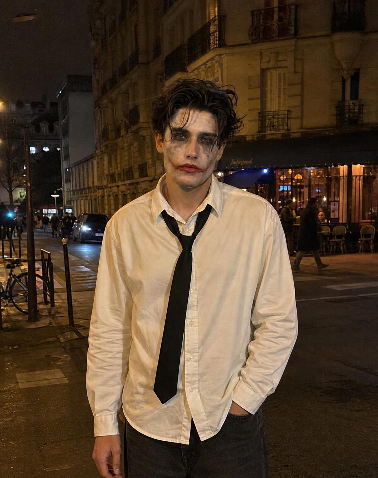 Create a photorealistic, natural Pinterest-style Halloween street photograph using the uploaded image ONLY as a facial identity reference.
Identity: Re-create the same person’s recognizable facial features, natural skin tone, and facial structure. Integrate their face naturally into the new photograph. Do not paste or overlay the reference face.
Halloween look: Realistic Joker-inspired makeup with imperfect white face paint, smudged black eye makeup with subtle streaks down the cheeks, and deep burgundy-red lipstick extending slightly beyond the lips. Keep the makeup believable, artistic, and hand-applied.
Outfit: Oversized, slightly wrinkled white button-up shirt, loosely hanging black tie, and dark baggy jeans.
Pose: Both hands completely empty. No microphone, no props, no objects in either hand. One arm hangs naturally beside the body while the other rests casually near the trouser pocket. Relaxed shoulders, natural posture, and a slightly mysterious expression. Avoid stiff or exaggerated posing.
Background: A real-looking European city street at night, elegant old buildings, wrought-iron balconies, illuminated storefronts, bicycles, passing cars, and warm amber streetlights.
Photography: Candid Halloween night-out photo taken casually by a friend on a smartphone. Natural flash mixed with ambient street lighting, subtle grain, realistic skin texture, slight imperfections, authentic shadows, muted colors, and effortless Pinterest aesthetics. The face, hair, neck, clothing, and background must share the same lighting, focus, camera perspective, and image texture.
Framing: Vertical 3:4 photograph, full head and upper body visible down to the upper thighs. Natural camera distance, slightly imperfect composition, no extreme close-up.
Avoid: Microphone, musical equipment, props, face cutout, pasted-face effect, mismatched lighting, artificial skin, excessive retouching, CGI, overly cinematic effects, stiff poses, distorted hands, and generic AI facial features.
Final result: A seamless, believable Halloween street photograph that looks genuinely captured in one camera shot, with the exact identity of the reference person and no artificial face-swap appearance.
