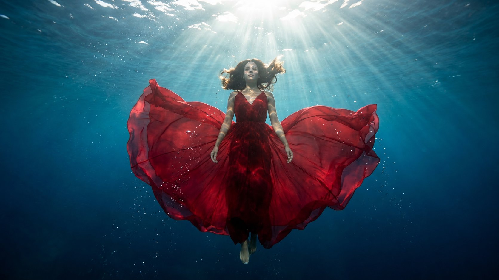 A woman in a flowing crimson gown suspended in turquoise water, backlit by sun rays breaking
through the
surface