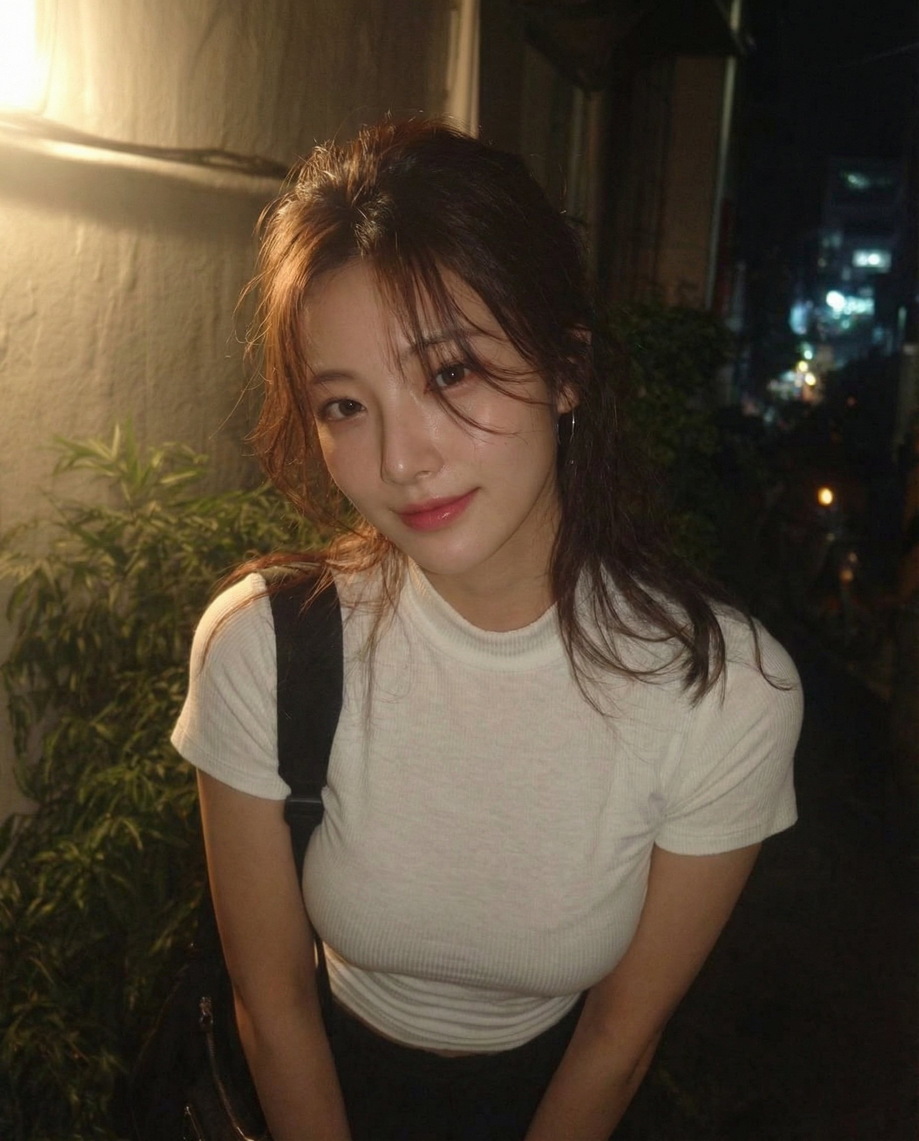 Photorealistic night-time portrait of a young East Asian woman standing outdoors in a narrow urban alley. She is framed from waist up, slightly leaning forward, looking directly at the camera with a soft, confident smile. She has long dark brown hair loosely tied back with natural strands falling around her face, clear fair skin with realistic texture, minimal makeup, and natural pink lips. She wears a fitted white short-sleeve ribbed t-shirt and dark pants, with a black shoulder bag strap visible. Warm streetlight illumination from the side creates soft highlights on her face and gentle shadows. Background shows dim alley walls, greenery, and distant city lights with subtle bokeh. Shallow depth of field, slight handheld feel, natural lens softness, low-light grain. No stylization, no beauty filters, no cinematic exaggeration. Looks like a real night photo taken casually, intimate and authentic.