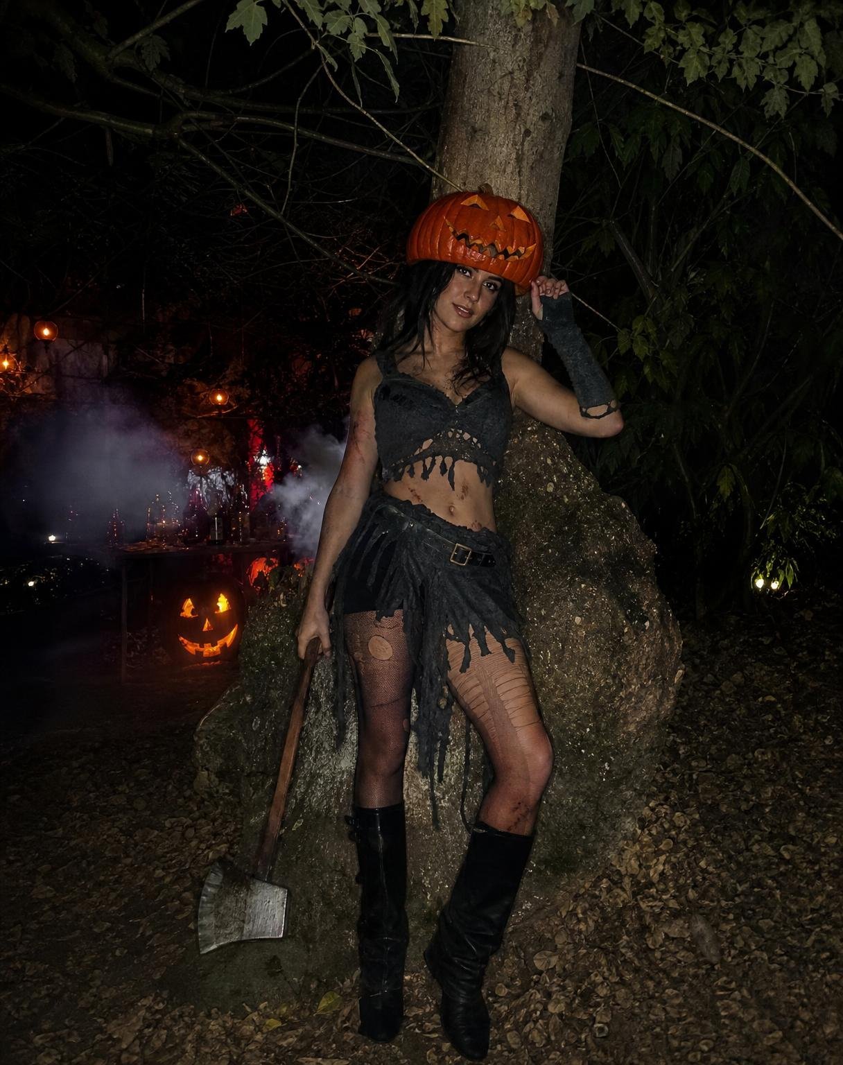Create an ultra-realistic, candid Halloween cosplay photograph of a young woman dressed as a dark pumpkin-headed slasher character. She wears a distressed, ripped Halloween outfit with torn fabric, rugged boots, and subtle dark, grungy styling. A carved orange pumpkin sits on top of her head as a creepy mask, with realistic carved eyes and a jagged mouth.
She is casually leaning against the trunk of a large, old tree in a spooky outdoor setting, holding a worn metal axe loosely in one hand. Her body language feels relaxed but unsettling, as if someone captured her unexpectedly during a Halloween night gathering. Fallen leaves, twisted tree branches, dim ambient light, and subtle Halloween atmosphere surround her.
Photography style: genuine smartphone snapshot, direct camera flash, realistic skin and fabric textures, natural shadows, imperfect framing, authentic nighttime atmosphere, slightly grainy image, Pinterest-worthy Halloween cosplay aesthetic. No professional photoshoot, no cinematic CGI, no overly polished AI look, no yellow color cast.
Face reference instructions: Use the provided reference image only for the woman’s identity and facial identity where visible. Do not copy the reference’s hairstyle, makeup, pose, expression, gaze, face angle, lighting, camera angle, or composition. The pumpkin headpiece must be positioned so it does not cover or replace her face; keep her real face visible beneath or alongside the pumpkin headpiece.
Square 1:1 composition. Full-body or three-quarter framing. Axe clearly visible, tree clearly visible, natural candid pose.