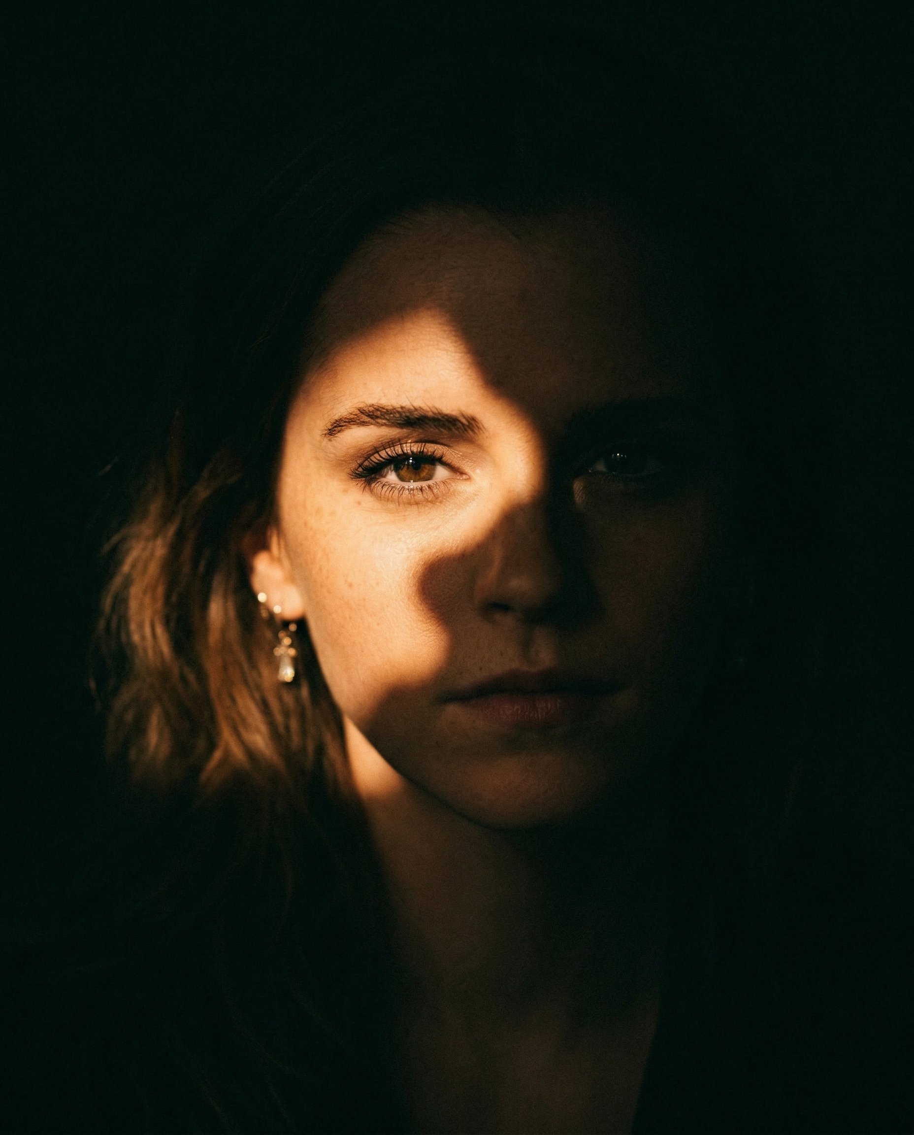 INPUT:
- Reference image of a person (must be used as identity base)

PROMPT:
Use the reference image as the exact identity base. Create a cinematic portrait with dramatic partial lighting, as if the subject is emerging from darkness with a strong, focused spotlight partially blocked by an object.

CORE LIGHTING (MOST IMPORTANT):
- Use a single hard spotlight coming from one side (slightly above eye level, angled)
- Simulate the light being **partially blocked by an object**, creating an irregular shadow cut across the face
- Only ONE side of the face should be illuminated (around one eye, cheek, and lips)
- The other side must fall into deep, pure black shadow
- The shadow edge must feel organic and imperfect (not a clean straight line)

LIGHT QUALITY:
- Strong, high contrast
- Bright highlight on skin where light hits
- Soft falloff into darkness
- No fill light — shadows must stay deep and rich

COMPOSITION:
- Slightly off-center framing (not perfectly symmetrical)
- Close portrait (head and shoulders)
- Natural angle, not stiff

FACE & EXPRESSION:
- Neutral or slightly intense expression
- Lips slightly parted (optional for cinematic feel)
- Eyes sharp and engaging

BACKGROUND:
- Completely dark or near-black
- No visible environment

STYLING:
- Hair softly lit on one side, fading into darkness on the other
- Optional subtle jewelry catching light (like earrings)
- Clothing dark, blending into shadows

COLOR GRADING:
- Warm cinematic tone on highlights (golden/orange tint)
- Deep crushed blacks
- Slight film-like contrast

TEXTURE:
- Add fine film grain
- Slight natural imperfection (not overly clean or digital)

EFFECTS:
- NO glow
- NO neon
- NO glitch
- NO overlays

DEPTH:
- Strong subject separation using light vs darkness only

MOOD:
- Cinematic, intimate, slightly mysterious
- Feels like a movie still or vintage film frame

IMPORTANT:
- Do NOT evenly light the face
- Do NOT center the light
- The asymmetry and shadow cut is the key visual hook