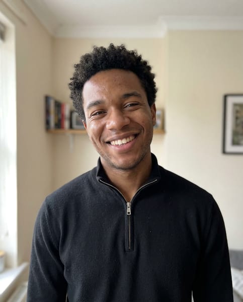 {
  "model": "NanoBanana",
  "task": "ultra realistic casual phone snapshot",
  "goal": "generate a highly natural, believable everyday portrait with zero studio feeling",
  "subject": {
    "person": "young man",
    "expression": "slightly asymmetric wide smile showing teeth, one lip corner raised a little higher, cheeks lifted with uneven natural tension, subtle eye squint, genuine candid joyful expression",
    "pose": "standing casually facing the camera, head tilted slightly a few degrees, shoulders relaxed, natural unstaged body language"
  },
  "appearance": {
    "hair": "short tight curly dark hair, natural shape, slightly imperfect everyday grooming",
    "skin": "visible pores, natural skin micro texture, subtle skin shine, slight tone variation, realistic facial imperfections, no retouching"
  },
  "clothing": {
    "top": "black quarter-zip sweater with silver zipper, simple casual everyday wear"
  },
  "environment": {
    "location": "ordinary indoor home environment",
    "background": "light indoor wall with subtle texture, faint shadow gradient, slight natural room depth, simple real-life setting",
    "context": "quick casual phone photo taken by a friend during a normal conversation"
  },
  "lighting": {
    "type": "natural mixed indoor light",
    "primary_source": "daylight coming from a nearby window on one side",
    "secondary_source": "soft ambient room light filling the opposite side",
    "effect": "slightly uneven illumination, soft natural shadow transitions, realistic exposure variation typical of phone photography"
  },
  "camera": {
    "device": "smartphone camera",
    "capture_style": "casual amateur phone snapshot",
    "lens_equivalent": "26mm smartphone lens",
    "framing": "head and shoulders portrait",
    "angle": "eye level",
    "focus": "natural phone autofocus on face",
    "depth_of_field": "natural smartphone depth",
    "artifacts": {
      "slight_noise": true,
      "subtle_compression": true,
      "minimal_lens_distortion": true,
      "slightly_compressed_dynamic_range": true,
      "minor_imperfection_in_sharpness": true
    }
  },
  "rendering": {
    "photorealism": "maximum",
    "style": "real everyday phone photography",
    "image_quality": "natural, candid, unpolished, believable",
    "detail": "high facial realism without looking processed or commercial"
  },
  "negative": [
    "studio portrait",
    "professional photoshoot",
    "beauty lighting",
    "perfect skin retouch",
    "plastic skin",
    "ai smile",
    "perfect symmetry",
    "cgi look",
    "cartoon style",
    "over smoothing",
    "commercial headshot",
    "over sharpened face",
    "harsh key light",
    "clean seamless backdrop",
    "fashion editorial look",
    "glamour portrait"
  ]
}