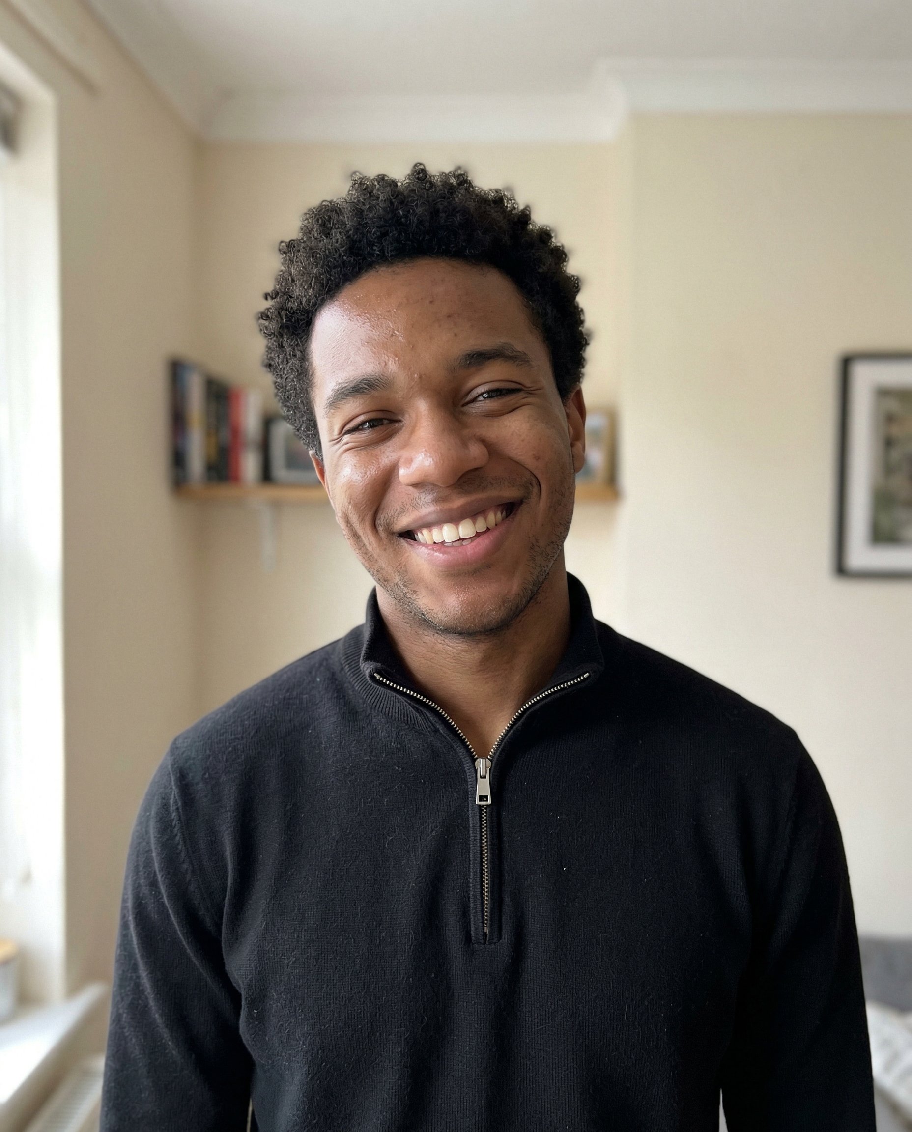 {
  "model": "NanoBanana",
  "task": "ultra realistic casual phone snapshot",
  "goal": "generate a highly natural, believable everyday portrait with zero studio feeling",
  "subject": {
    "person": "young man",
    "expression": "slightly asymmetric wide smile showing teeth, one lip corner raised a little higher, cheeks lifted with uneven natural tension, subtle eye squint, genuine candid joyful expression",
    "pose": "standing casually facing the camera, head tilted slightly a few degrees, shoulders relaxed, natural unstaged body language"
  },
  "appearance": {
    "hair": "short tight curly dark hair, natural shape, slightly imperfect everyday grooming",
    "skin": "visible pores, natural skin micro texture, subtle skin shine, slight tone variation, realistic facial imperfections, no retouching"
  },
  "clothing": {
    "top": "black quarter-zip sweater with silver zipper, simple casual everyday wear"
  },
  "environment": {
    "location": "ordinary indoor home environment",
    "background": "light indoor wall with subtle texture, faint shadow gradient, slight natural room depth, simple real-life setting",
    "context": "quick casual phone photo taken by a friend during a normal conversation"
  },
  "lighting": {
    "type": "natural mixed indoor light",
    "primary_source": "daylight coming from a nearby window on one side",
    "secondary_source": "soft ambient room light filling the opposite side",
    "effect": "slightly uneven illumination, soft natural shadow transitions, realistic exposure variation typical of phone photography"
  },
  "camera": {
    "device": "smartphone camera",
    "capture_style": "casual amateur phone snapshot",
    "lens_equivalent": "26mm smartphone lens",
    "framing": "head and shoulders portrait",
    "angle": "eye level",
    "focus": "natural phone autofocus on face",
    "depth_of_field": "natural smartphone depth",
    "artifacts": {
      "slight_noise": true,
      "subtle_compression": true,
      "minimal_lens_distortion": true,
      "slightly_compressed_dynamic_range": true,
      "minor_imperfection_in_sharpness": true
    }
  },
  "rendering": {
    "photorealism": "maximum",
    "style": "real everyday phone photography",
    "image_quality": "natural, candid, unpolished, believable",
    "detail": "high facial realism without looking processed or commercial"
  },
  "negative": [
    "studio portrait",
    "professional photoshoot",
    "beauty lighting",
    "perfect skin retouch",
    "plastic skin",
    "ai smile",
    "perfect symmetry",
    "cgi look",
    "cartoon style",
    "over smoothing",
    "commercial headshot",
    "over sharpened face",
    "harsh key light",
    "clean seamless backdrop",
    "fashion editorial look",
    "glamour portrait"
  ]
}