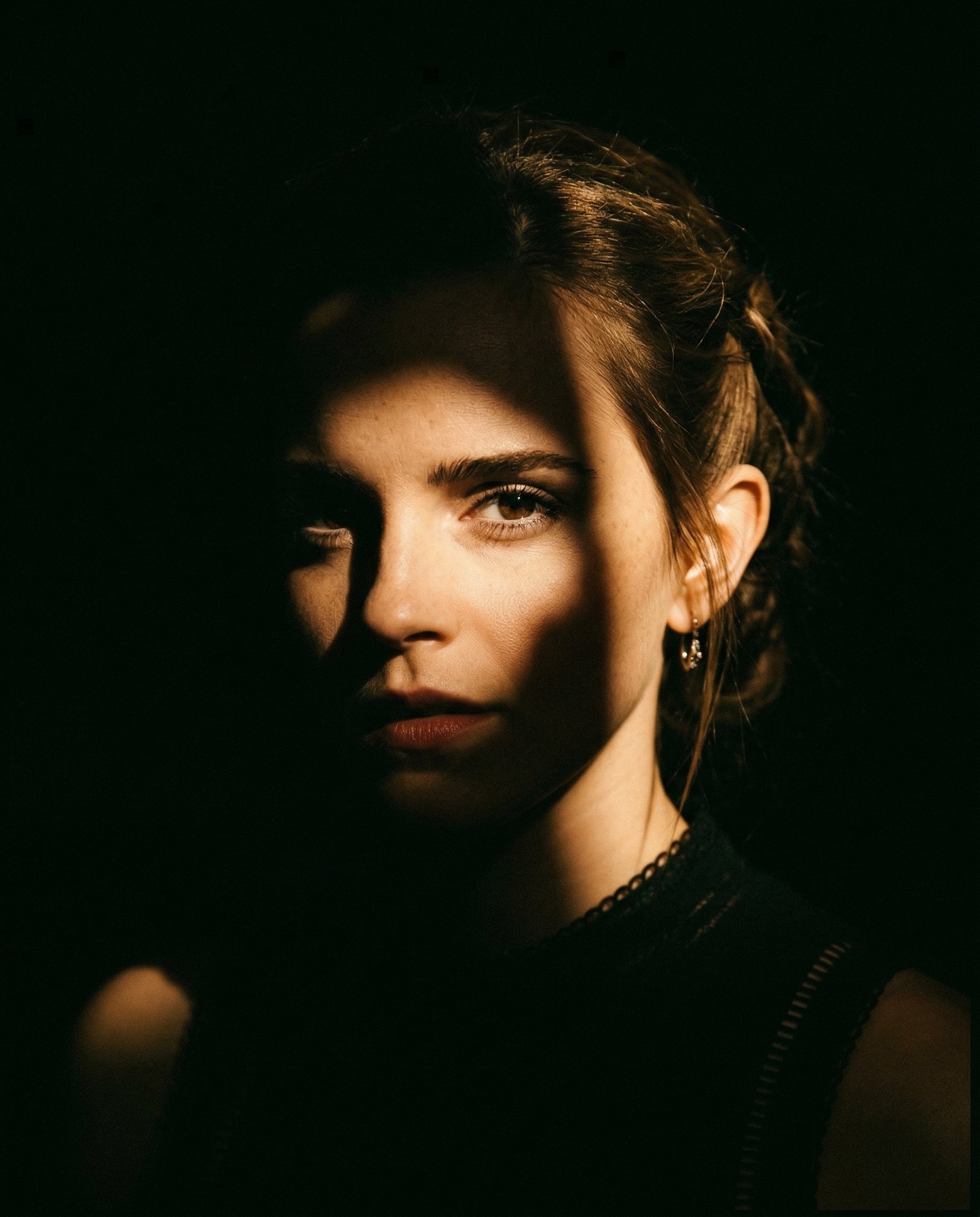 INPUT:
- Reference image of a person (must be used as identity base)

PROMPT:
Use the reference image as the exact identity base. Create a cinematic portrait with dramatic partial lighting, as if the subject is emerging from darkness with a strong, focused spotlight partially blocked by an object.

CORE LIGHTING (MOST IMPORTANT):
- Use a single hard spotlight coming from one side (slightly above eye level, angled)
- Simulate the light being **partially blocked by an object**, creating an irregular shadow cut across the face
- Only ONE side of the face should be illuminated (around one eye, cheek, and lips)
- The other side must fall into deep, pure black shadow
- The shadow edge must feel organic and imperfect (not a clean straight line)

LIGHT QUALITY:
- Strong, high contrast
- Bright highlight on skin where light hits
- Soft falloff into darkness
- No fill light — shadows must stay deep and rich

COMPOSITION:
- Slightly off-center framing (not perfectly symmetrical)
- Close portrait (head and shoulders)
- Natural angle, not stiff

FACE & EXPRESSION:
- Neutral or slightly intense expression
- Lips slightly parted (optional for cinematic feel)
- Eyes sharp and engaging

BACKGROUND:
- Completely dark or near-black
- No visible environment

STYLING:
- Hair softly lit on one side, fading into darkness on the other
- Optional subtle jewelry catching light (like earrings)
- Clothing dark, blending into shadows

COLOR GRADING:
- Warm cinematic tone on highlights (golden/orange tint)
- Deep crushed blacks
- Slight film-like contrast

TEXTURE:
- Add fine film grain
- Slight natural imperfection (not overly clean or digital)

EFFECTS:
- NO glow
- NO neon
- NO glitch
- NO overlays

DEPTH:
- Strong subject separation using light vs darkness only

MOOD:
- Cinematic, intimate, slightly mysterious
- Feels like a movie still or vintage film frame

IMPORTANT:
- Do NOT evenly light the face
- Do NOT center the light
- The asymmetry and shadow cut is the key visual hook