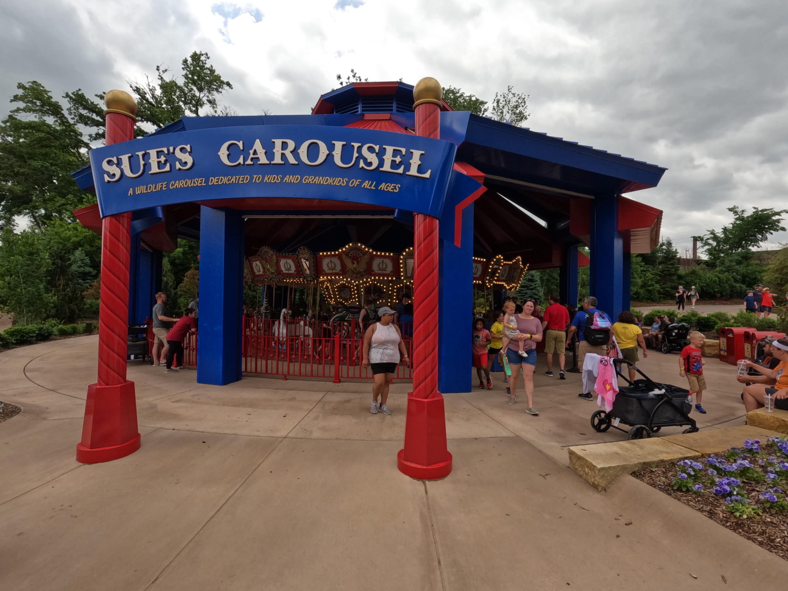 Sue's Carousel entrance archway with the carousel running in the background. Patrons are standing around the carousel watching.  