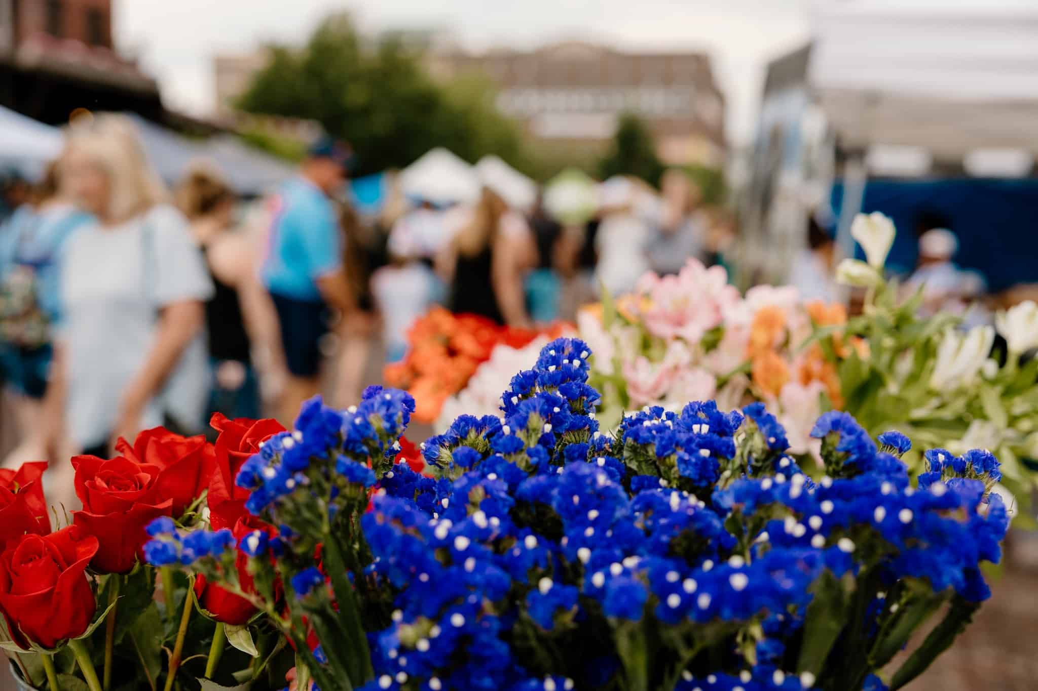 Flowers in the foreground with farmers market patrons browsing stalls in the background. 