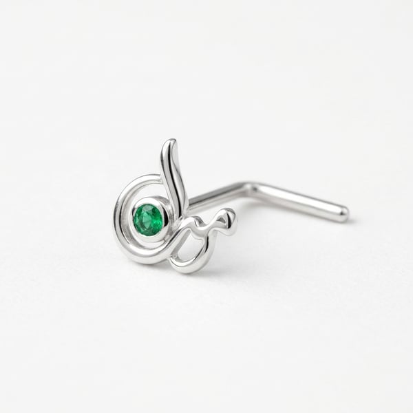 Macro shot of the Ijlaal sterling silver nose stud featuring a smooth Arabic calligraphy line with a tiny bezel-set round green emerald on a plain white surface.