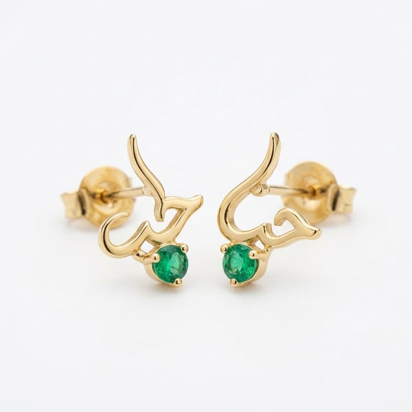 Pair of Zayan 18K gold stud earrings featuring a subtle Arabic calligraphy line and natural round emeralds on a white background.