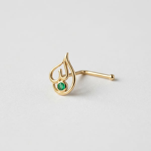 Studio shot of Rayan 18K gold nose pin with Arabic calligraphy motif and a 2 mm round natural emerald on a white surface.