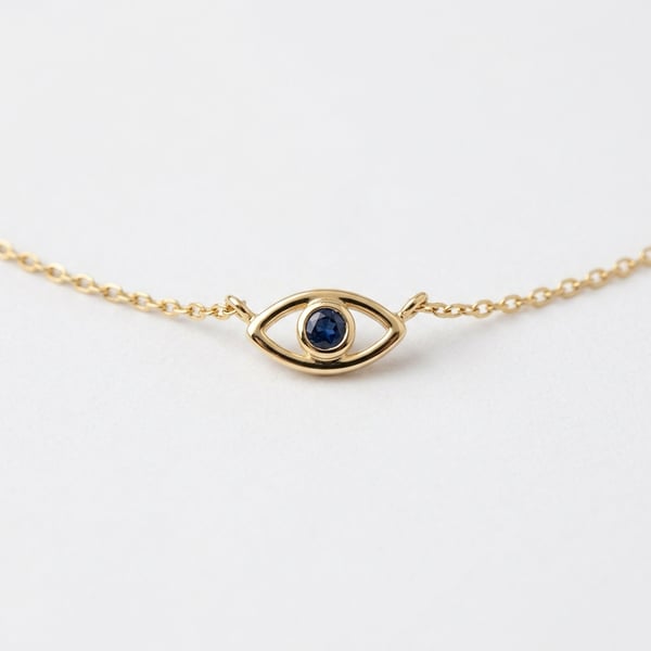 Studio shot of 18K gold Sayida evil eye necklace with a bezel-set round blue sapphire center stone on a white background.