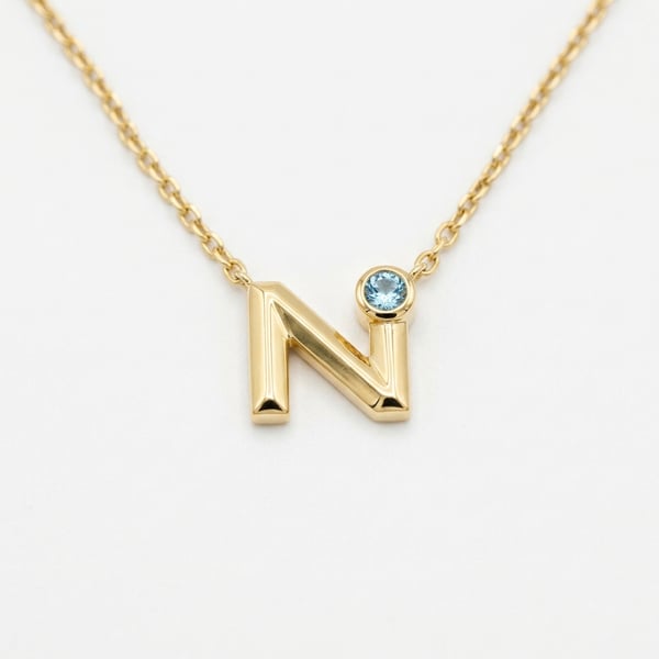 18K gold geometric letter N pendant with a bezel-set round aquamarine on a fine gold chain.