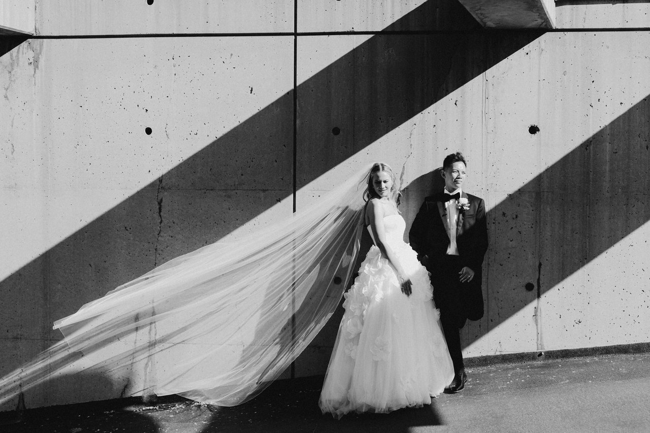 Editorial black-and-white wedding portrait — bride and groom against a concrete wall, veil caught mid-motion in the wind