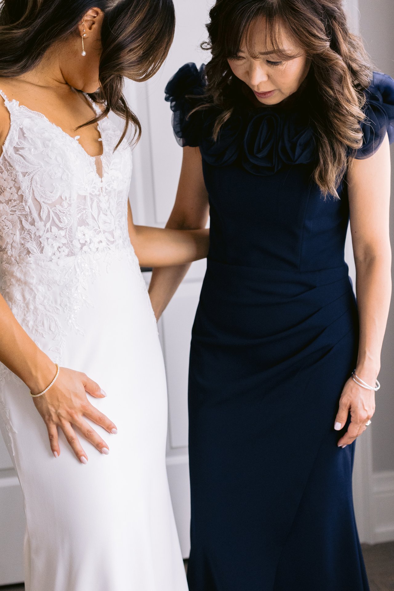 Bride and her mother standing close together during getting-ready, mother's hand on the bride's back, editorial-cropped frame in soft window light