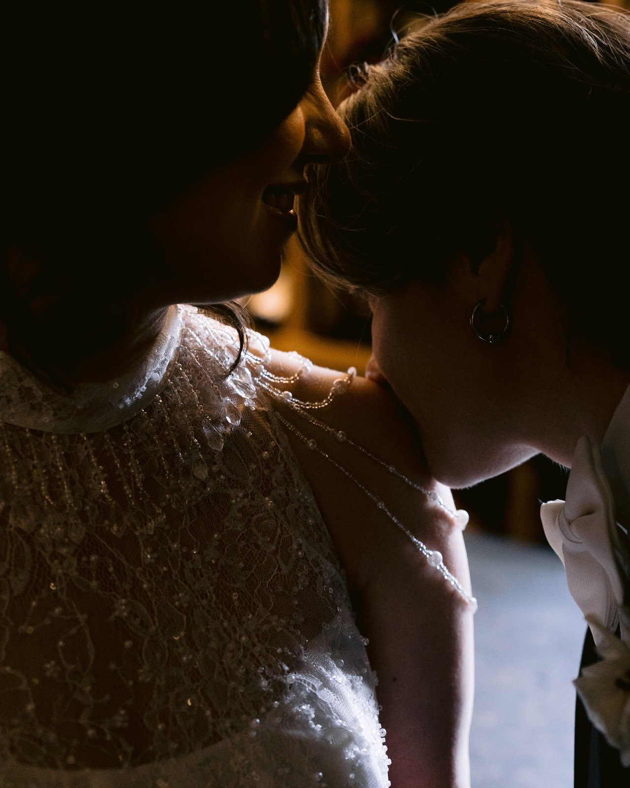 Intimate candid wedding moment — bride in beaded lace dress shares a quiet exchange with a loved one, soft ambient light