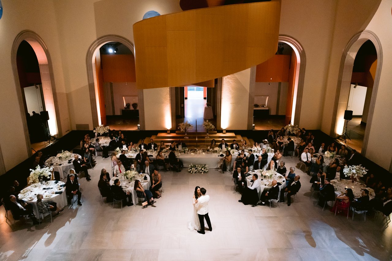 First dance at the Art Gallery of Ontario (AGO) — couple on the empty dance floor surrounded by guests at round tables, Frank Gehry wooden curves above
