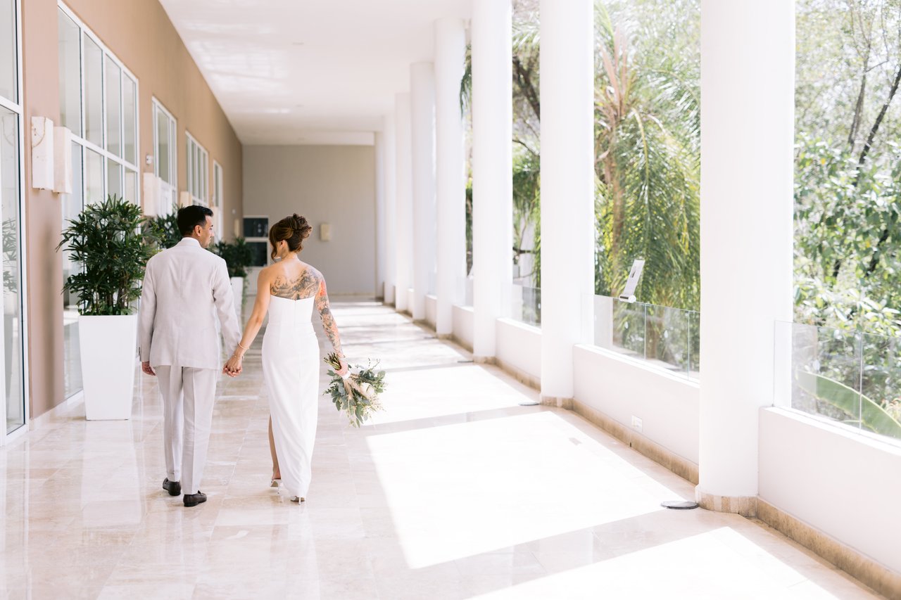 Bride and groom walking hand in hand down a marble destination-wedding corridor — tropical greenery through the windows, white columns receding into soft daylight