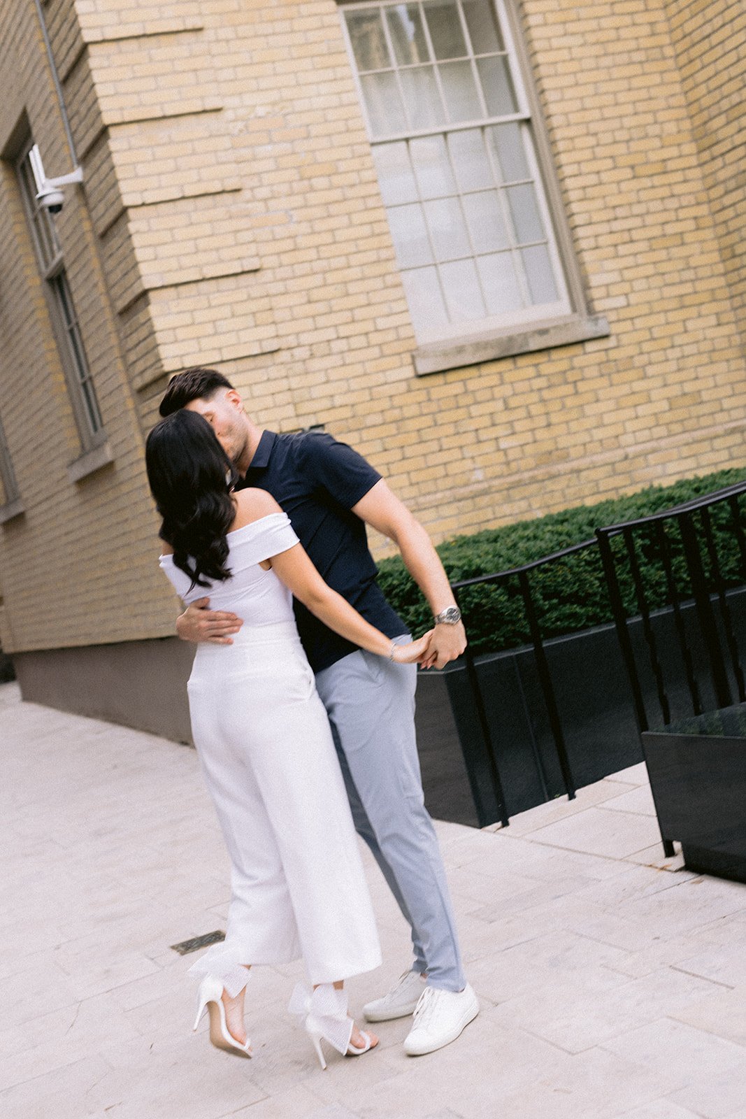 Engagement-session embrace on a Toronto sidewalk — bride in white off-shoulder jumpsuit, groom in navy polo, yellow-brick architecture and soft afternoon light