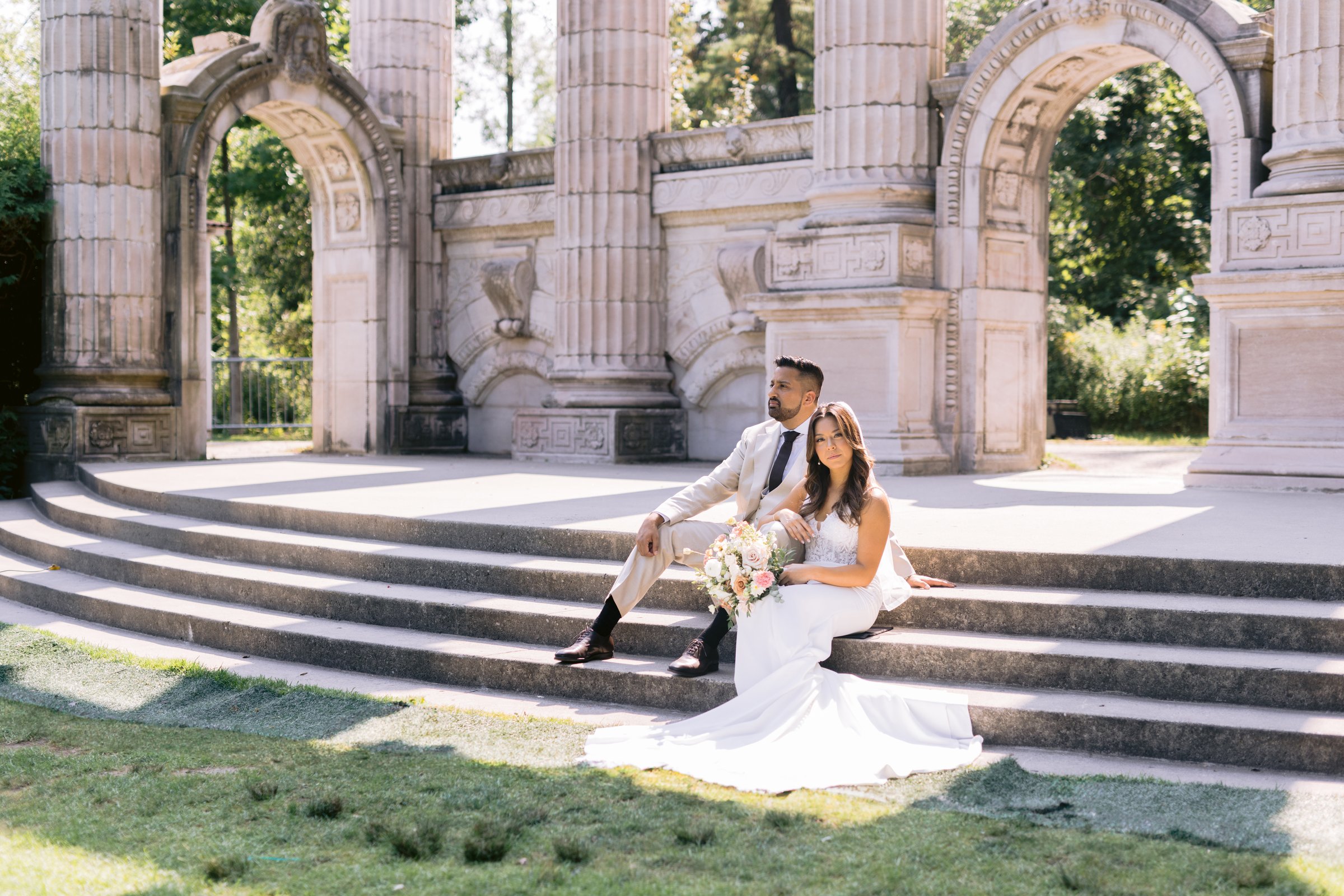 Midori and Adil seated on the stone amphitheatre steps at Guildwood Park, the Greek-style arches and columns behind them