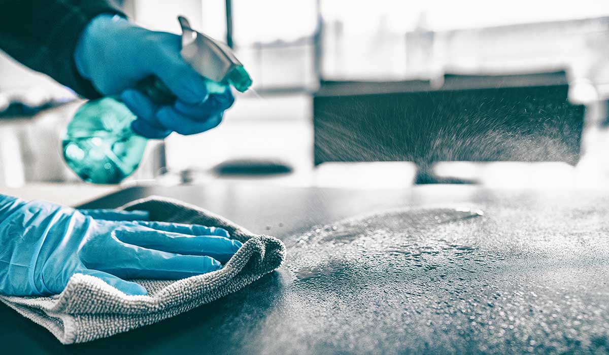 Botella pulverizando alcohol isopropílico, mientras con la otra mano usa una bayeta para limpiar la superficie.