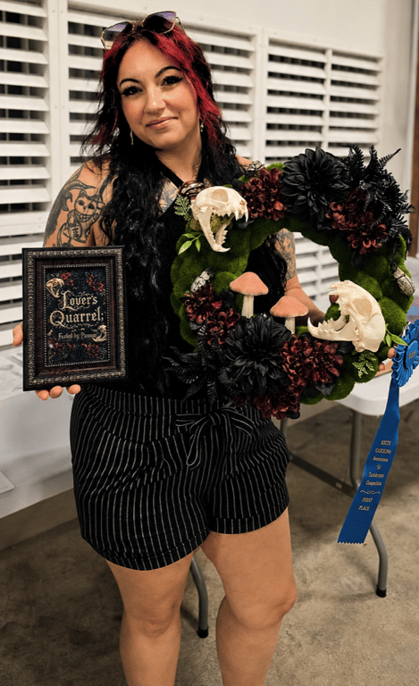 Picture of a tattooed cutie, female, black hair with red bangs, sunglasses, holding taxidermy art wreath