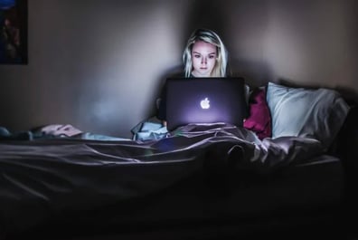 Lady sitting in bed in a dark room on her laptop with light highlighting her face
