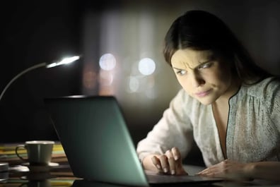 Mujer mirando confundida una computadora portátil debajo de una pequeña lámpara de escritorio que representa a alguien que se pregunta cómo encontrar quién está detrás de una cuenta falsa de fakebook.