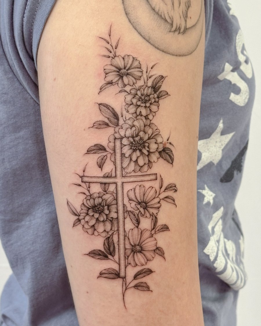 A delicate black and grey cross with flowers tattoo on the upper arm, featuring fine line work and floral details — by Melanie Benoit, los angeles