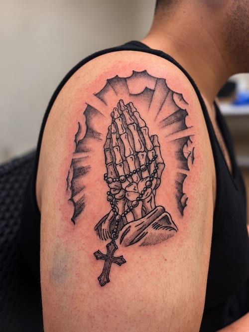 A bold black and grey tattoo of praying hands with a rosary and cross on the upper arm, featuring spiritual and religious themes — by HEAVY INK, los angeles