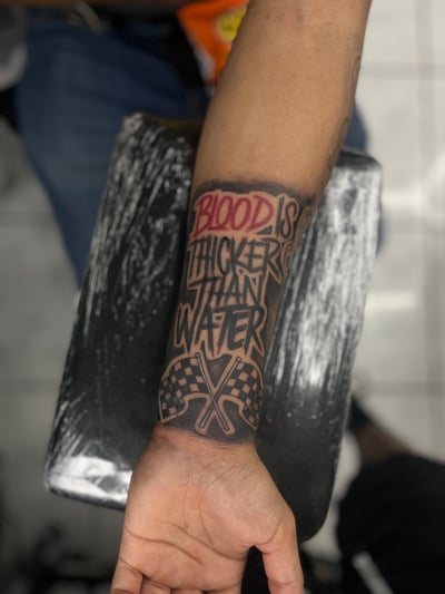 A bold lettering tattoo on the forearm with the phrase 'Blood is Thicker Than Water' in black and red ink, featuring checkered flags at the bottom — by Mr._Folks, los angeles
