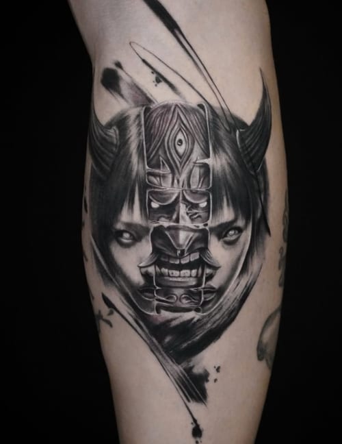 A black and grey surrealistic tattoo on the calf, featuring a demon mask overlaying a woman's face with intricate details and bold blackwork shading — by Jian Liang, los angeles