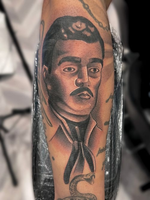 A black and grey Chicano style portrait tattoo on the upper arm, depicting a man with detailed shading and strong facial features — by Josue Bravo, los angeles