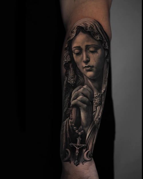 A realistic black and grey tattoo of a praying woman holding a rosary, with a cross, on the forearm — by Vladimir Kozachenko, los angeles