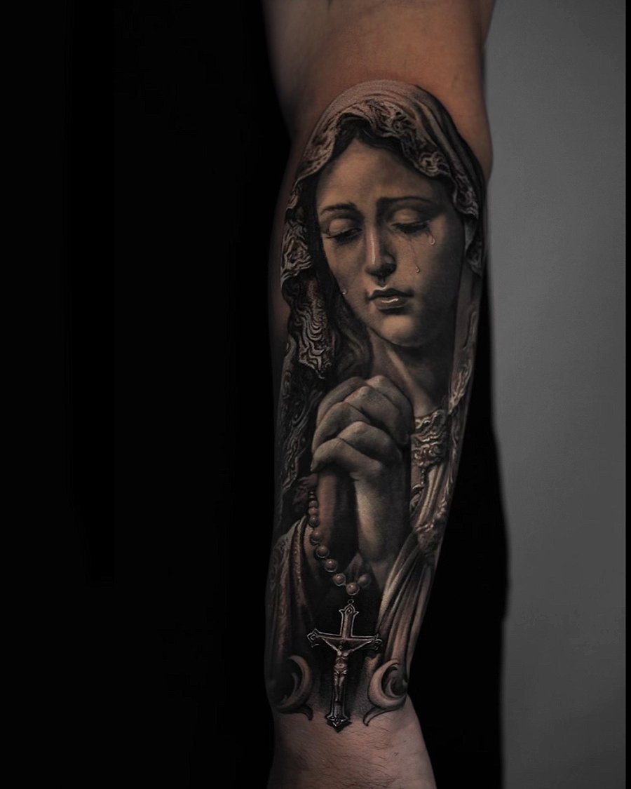 A realistic black and grey tattoo of a praying woman holding a rosary, with a cross, on the forearm — by Vladimir Kozachenko, los angeles