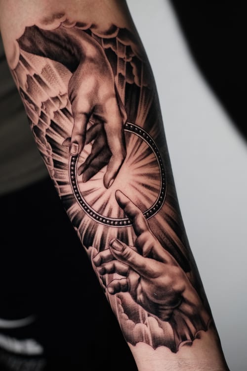 A black and grey realism tattoo on the forearm depicting two hands reaching towards each other with intricate shading and detail — by Johnny Ayala, los angeles