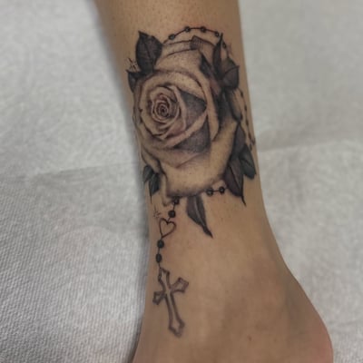 A black and grey rose with a rosary and cross on the ankle, featuring fine line details — by Maddie Ramirez, los angeles