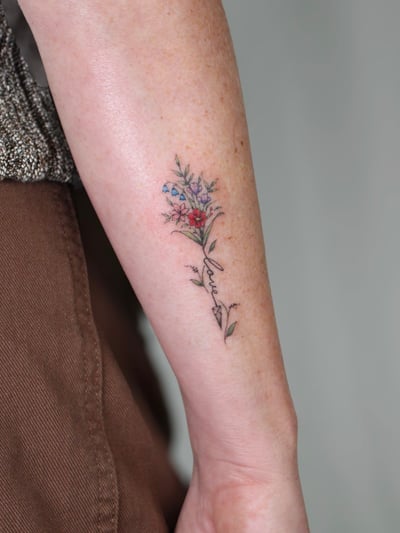 A delicate fine line floral bouquet with script on the forearm, featuring small colorful flowers — by Melanie Benoit, los angeles